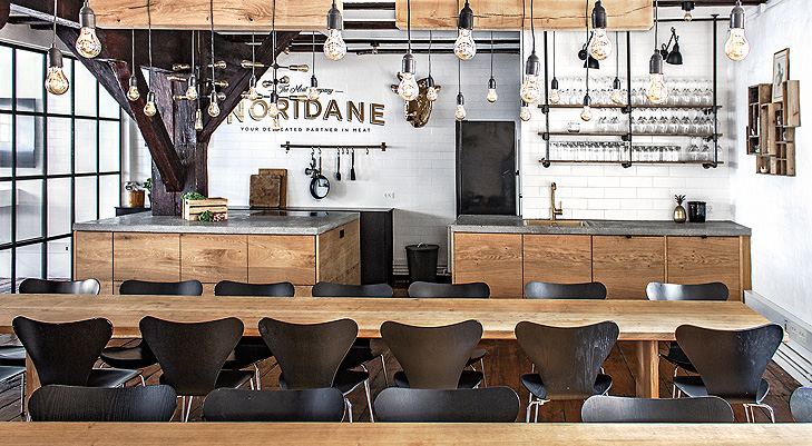 Overview of the interiors of a canteen med massive raw oak wood tables and balck wooden chairs. In the ceiling there is hagning some decorative oak beams with light bulps in black wires.